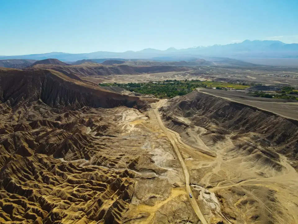 Vista aérea de un camino de tierra entre quebradas del desierto que baja hacia un oasis verde con cordillera al fondo