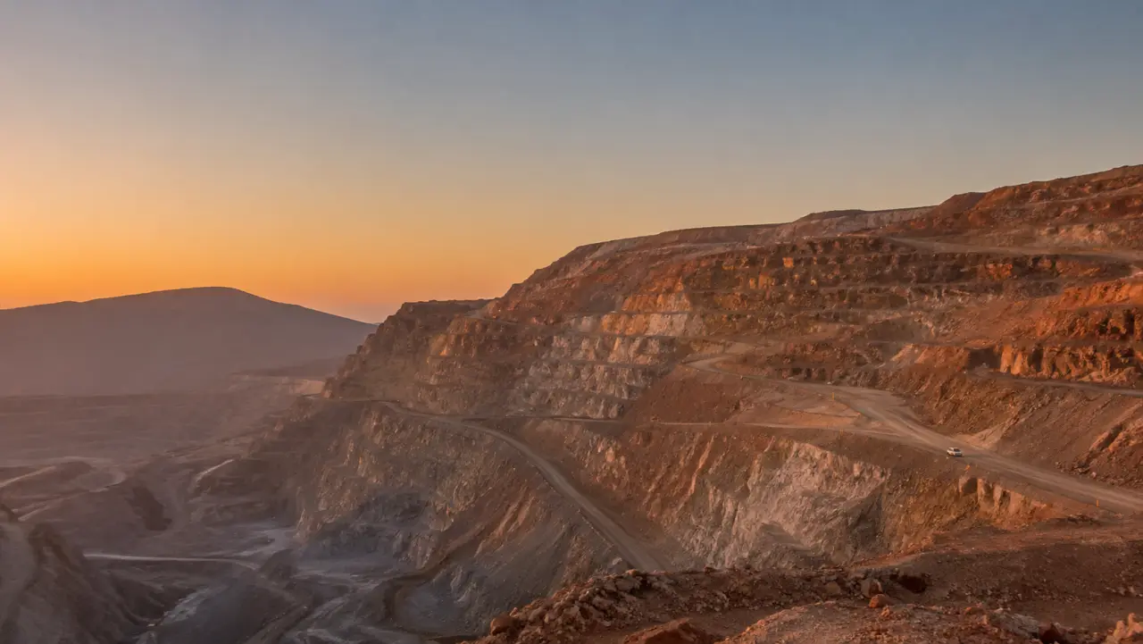 Faena minera a rajo abierto en el desierto al atardecer, con bancos escalonados y cerros al fondo