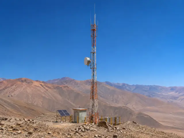 Torre de radio con antenas y un plato de enlace en la cima de un cerro, con caseta, panel solar y cerco, sobre un valle del desierto