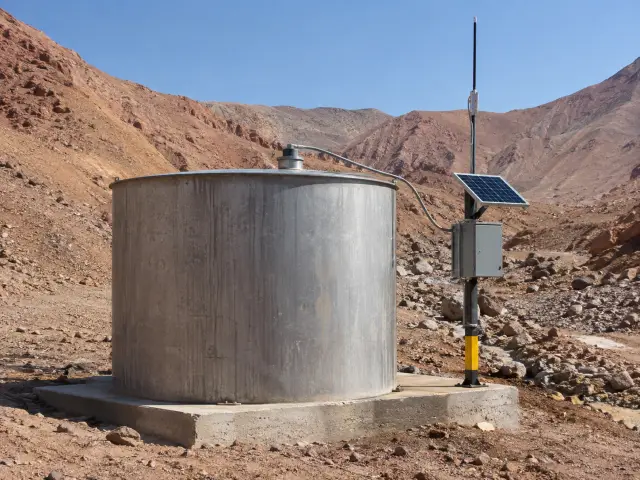 Estanque cilíndrico en una quebrada del desierto con sensor de nivel en la tapa y un poste con gabinete, panel solar pequeño y antena
